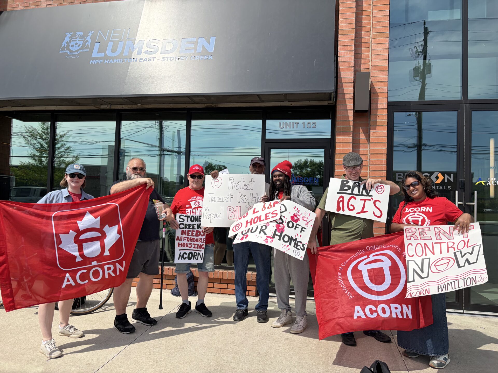 Group of protesters holding signs about tenant rights and rent control, with red ACORN banners outside a brick storefront.