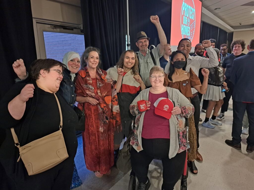 Group of diverse people posing with fists raised in a line at an event, some smiling, in front of a stage screen that reads 'Protect Our Homes'.