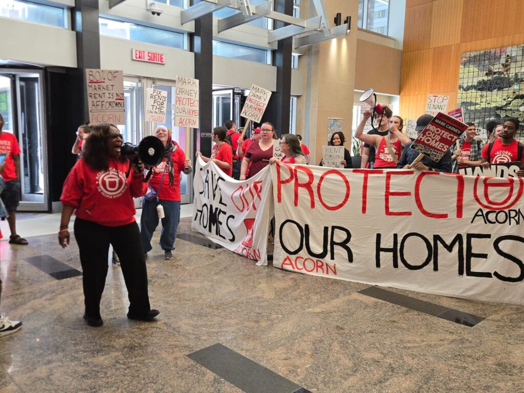 Protesters in red shirts hold a large banner reading 'PROTECT OUR HOMES' inside a building lobby, with signs about housing.
