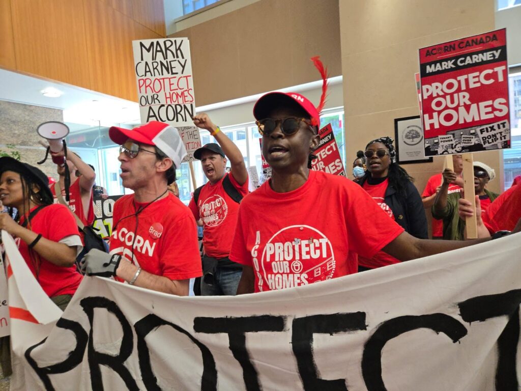 Group of protesters in red shirts marching indoors, holding a large banner and signs that read 'Protect Our Homes' and 'Mark Carney' inscriptions.