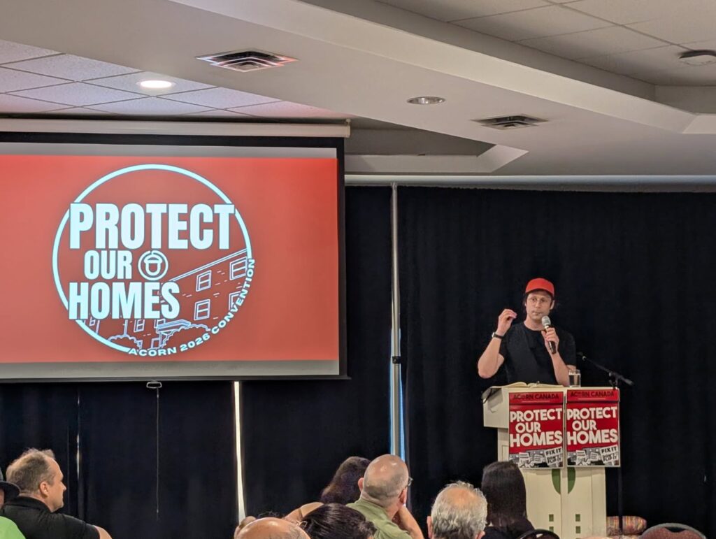 Speaker at a podium presenting a 'Protect Our Homes' banner; large red screen shows the same logo with a map outline in the background (ACORN Canada event).