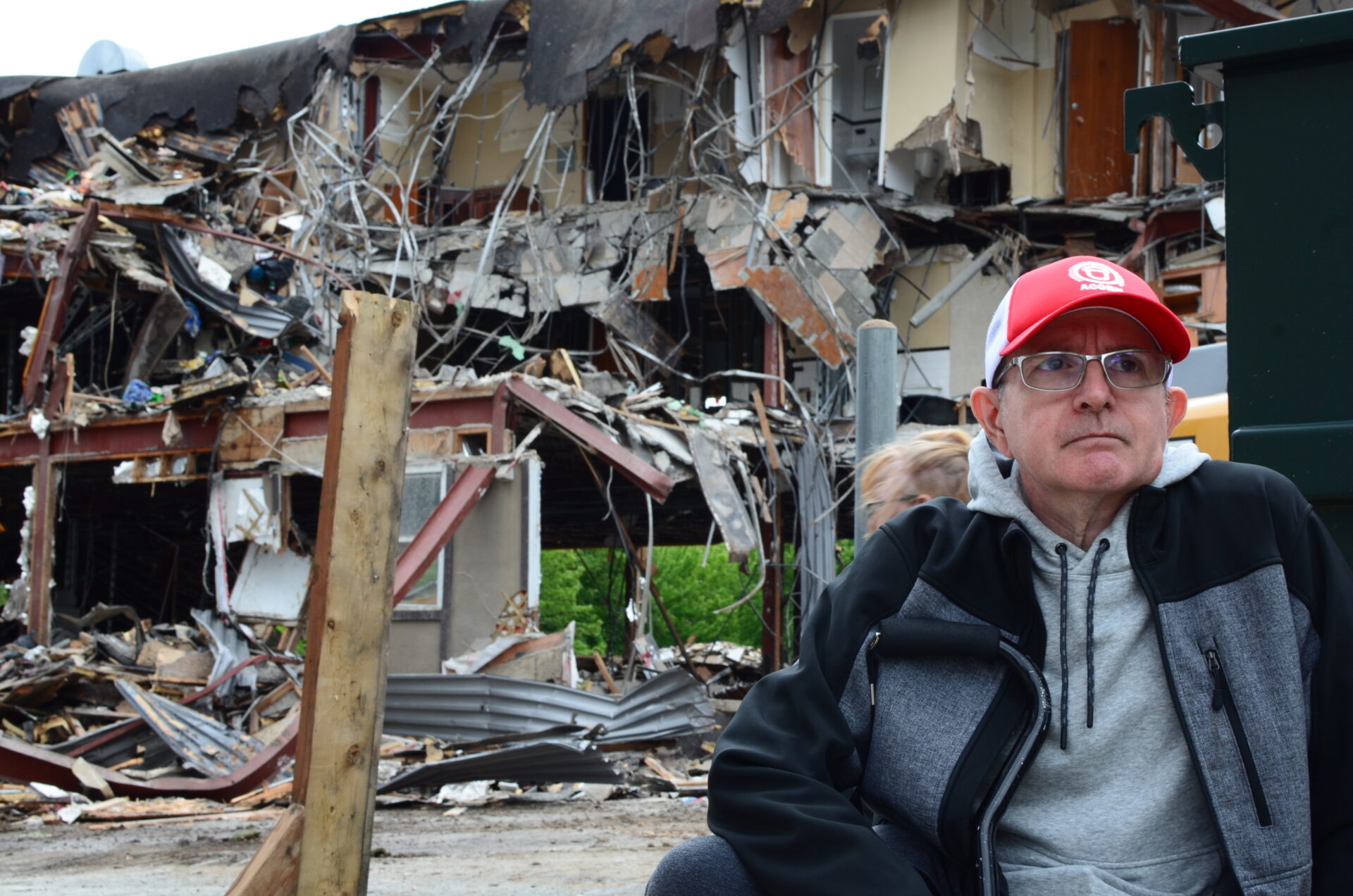 Man in glasses and a red cap stands beside a collapsed building with rubble and tangled metal.