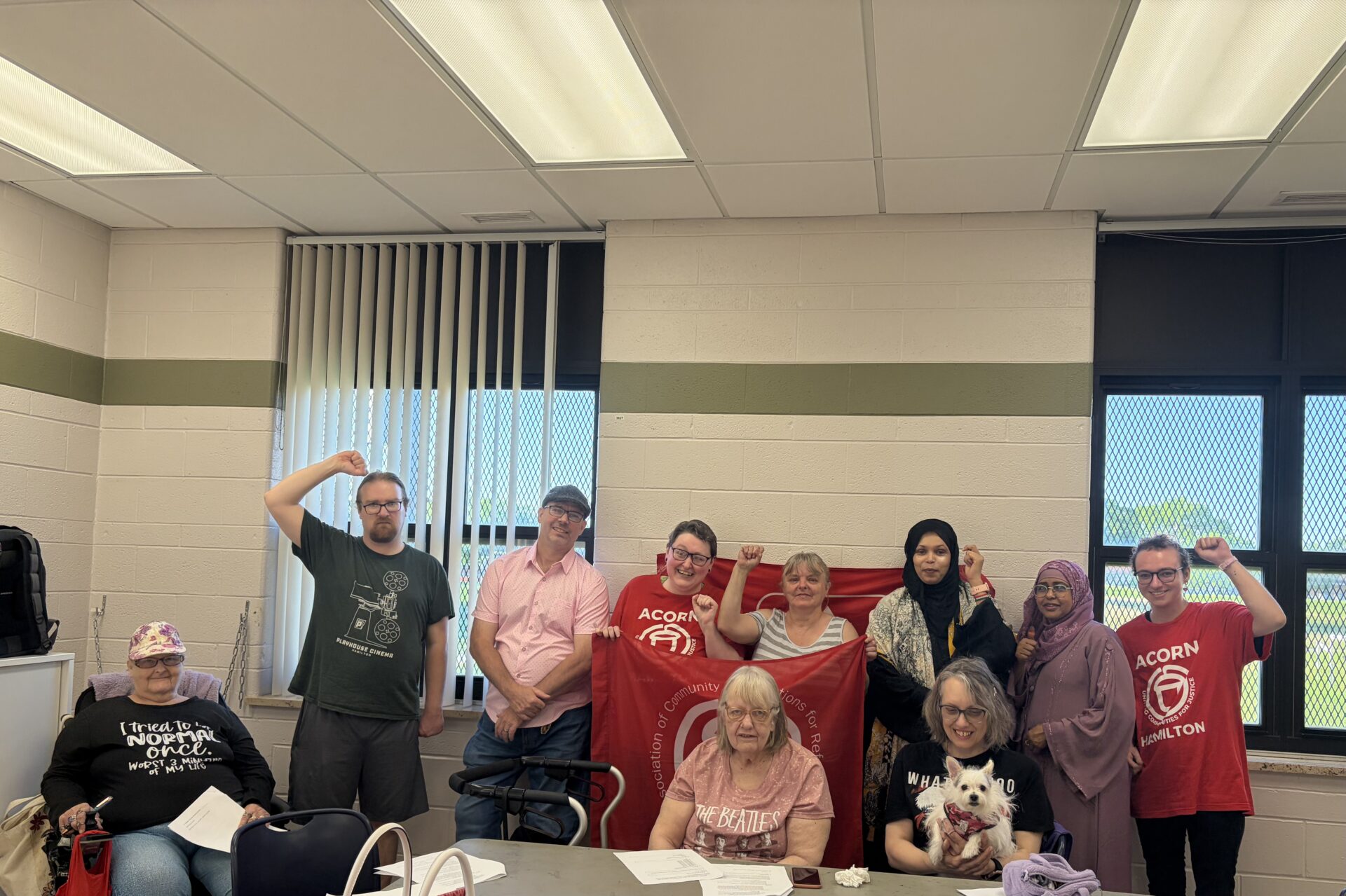 Group photo of diverse adults in a room, holding red banners and posing for the camera at a community event.