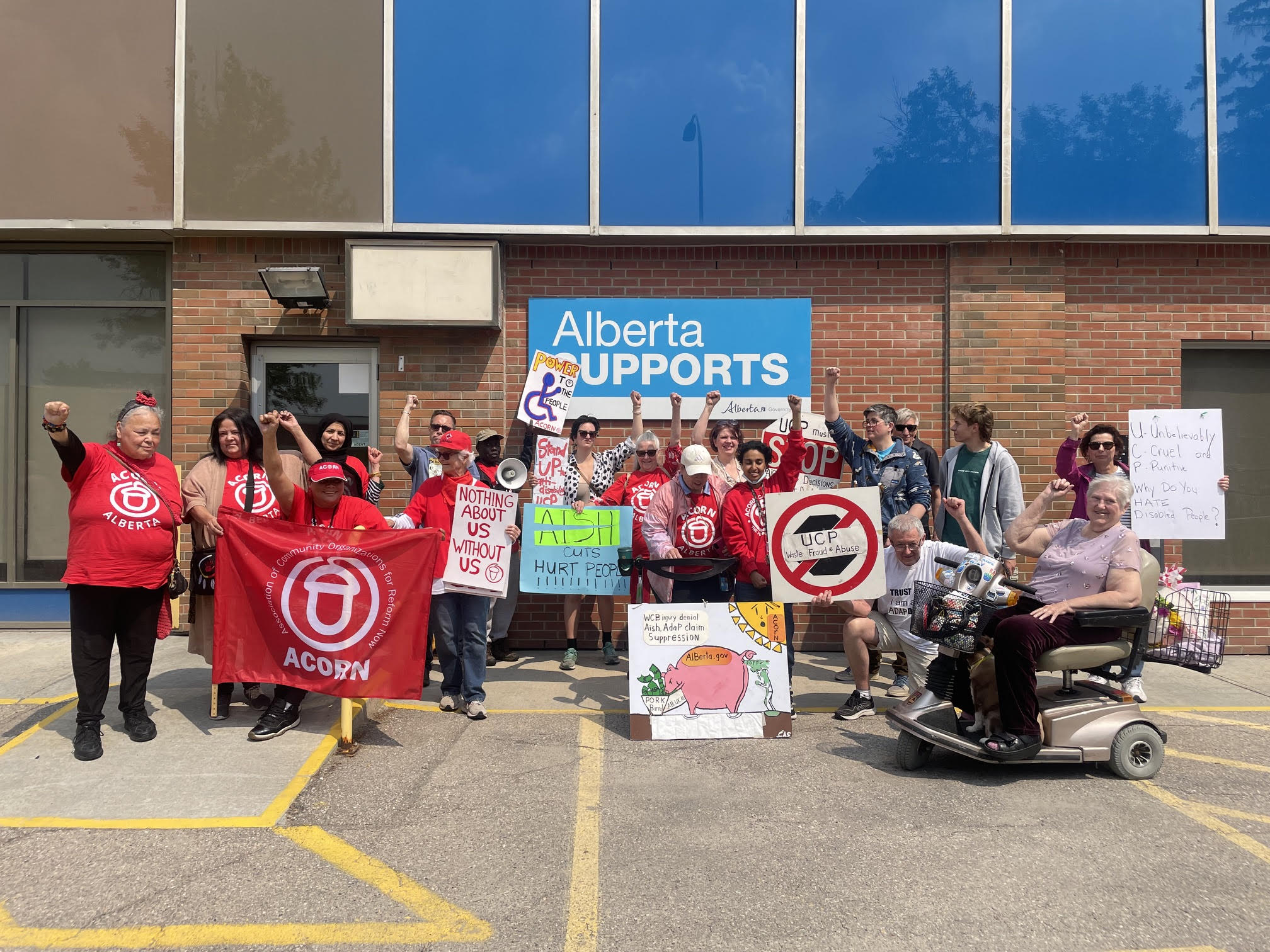Group of protesters outside a brick building holding signs and a blue Alberta Supports banner.