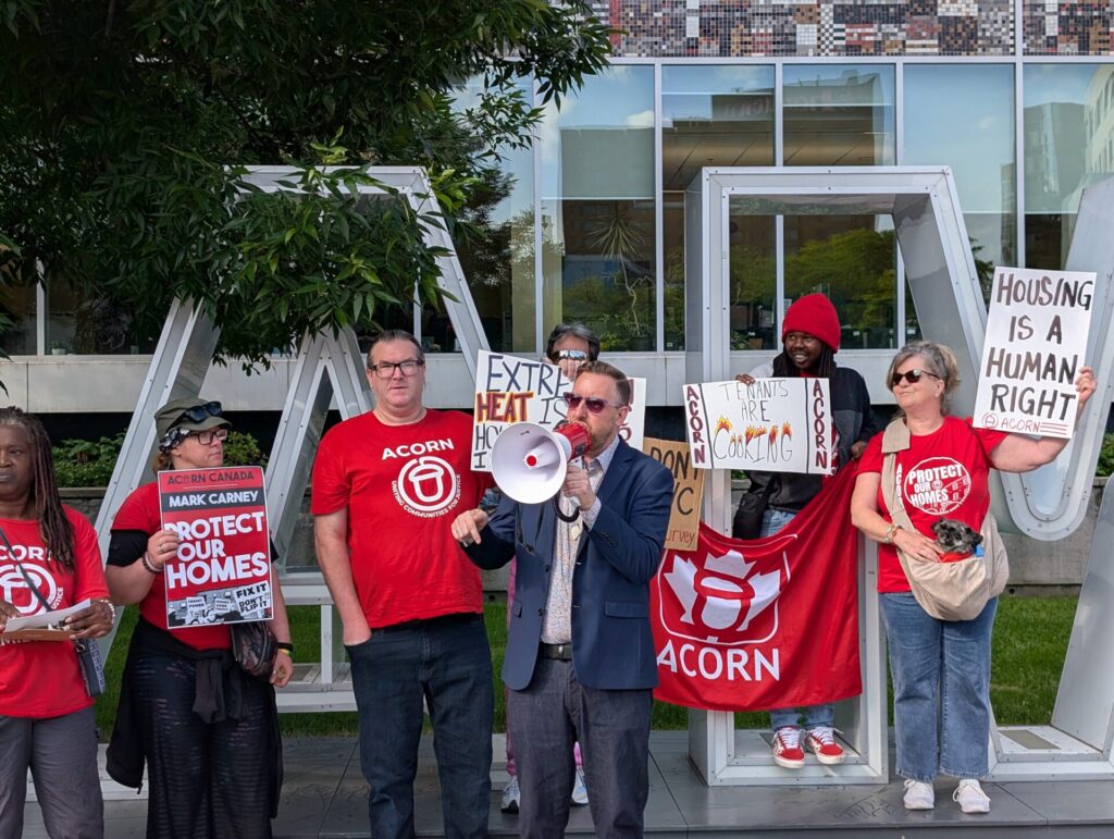 Group of protesters in red shirts holding signs about housing rights outside a modern glass building; sign reads 'Housing is a Human Right.'