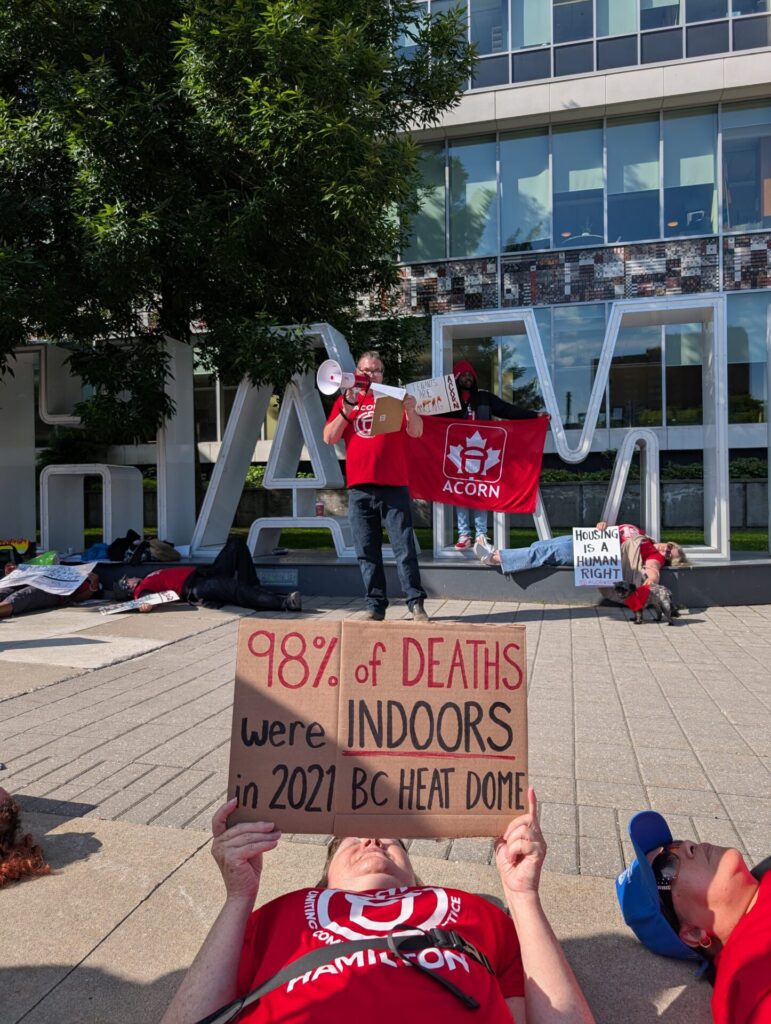 Protest scene outside a modern building: people in red shirts hold a red Acorn banner and signs; a foreground protester reads '98% of deaths were indoors' on cardboard.