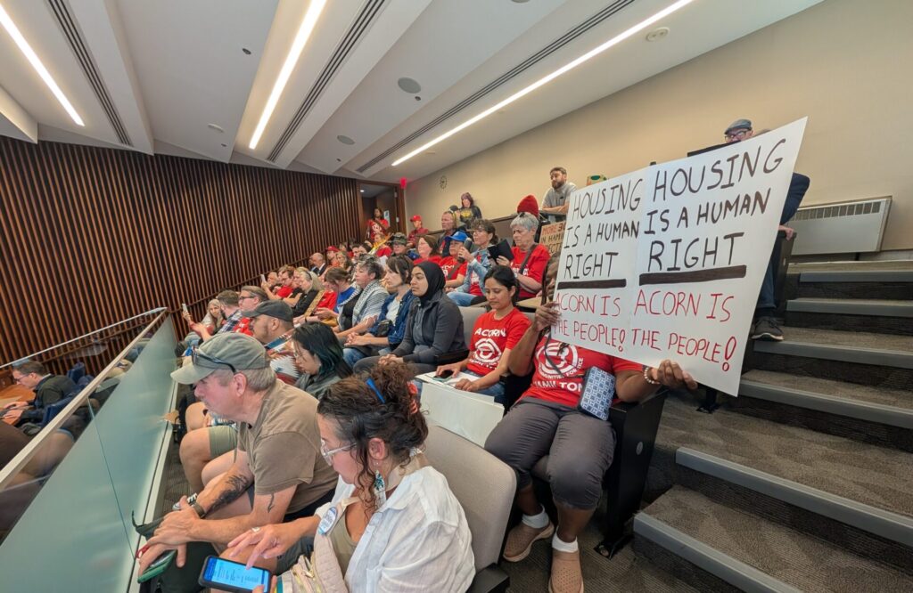 Crowd of people in a lecture hall holding signs that read 'Housing is a human right' during a protest. Women and men sit in rows wearing red shirts, some using phones or tablets.