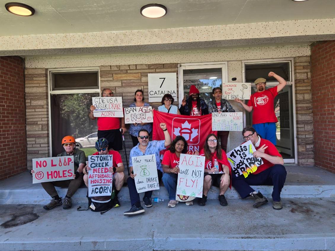 Group of protesters posing on the steps of a brick building, holding handmade signs calling for affordable housing and rent reform, with a red banner in the center.