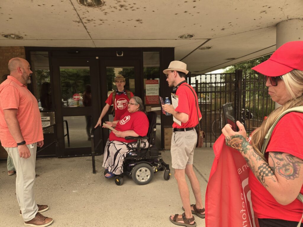 People stand outside a building entrance; a man in a pink shirt talks to a woman in a wheelchair while others in red shirts look on inside the doorway area.