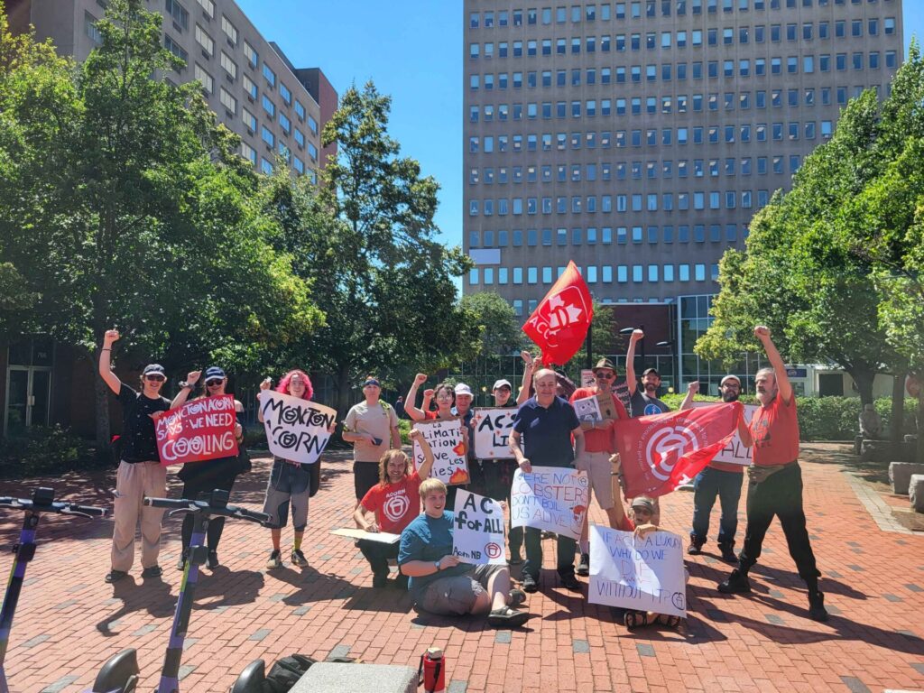 Group of diverse protesters outdoors in a city plaza, holding handmade signs and red banners, with tall office buildings and green trees in the background.