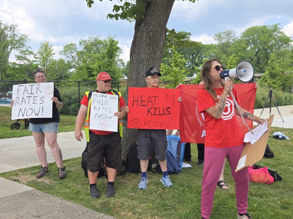 Group of protesters holding signs about fair rates, including 'FAIR RATES NOW' and 'HEAT KILLS ACORN', near a tree with a megaphone speaker on the right.