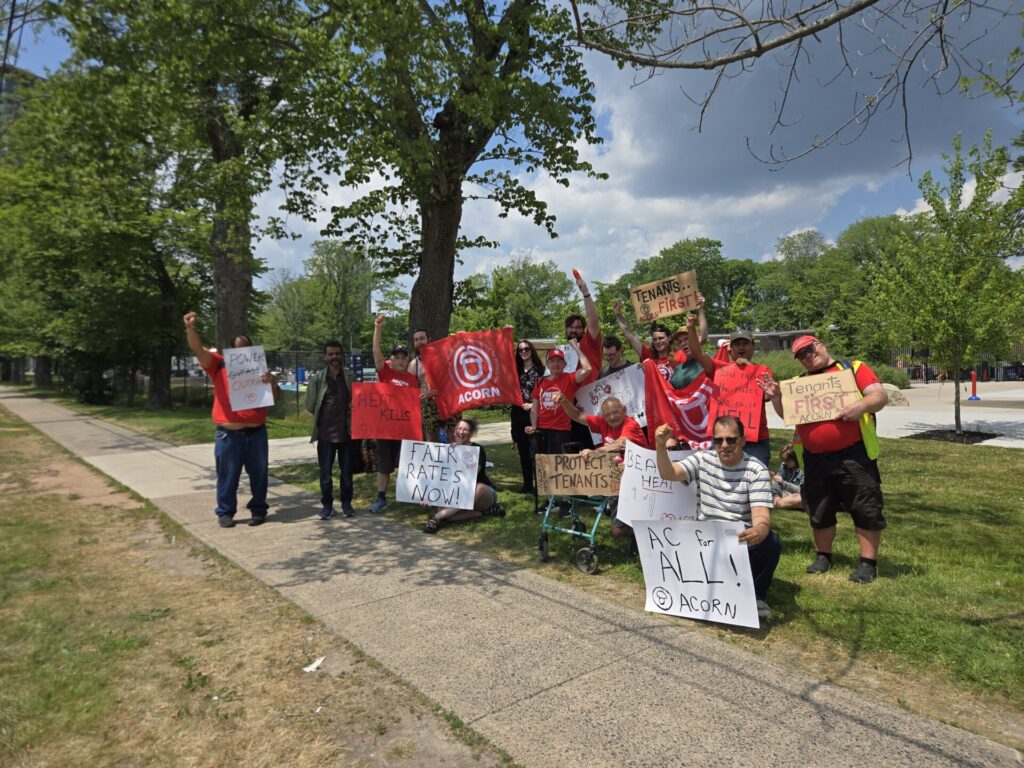 Group of protesters wearing red shirts holding signs and banners on a sunny street, some signs read Tenants First and ACORN.