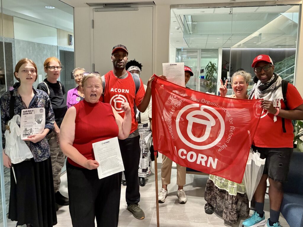 Diverse group indoors posing with a red ACORN banner and pamphlets, two people flashing peace signs in front of a glass doorway.