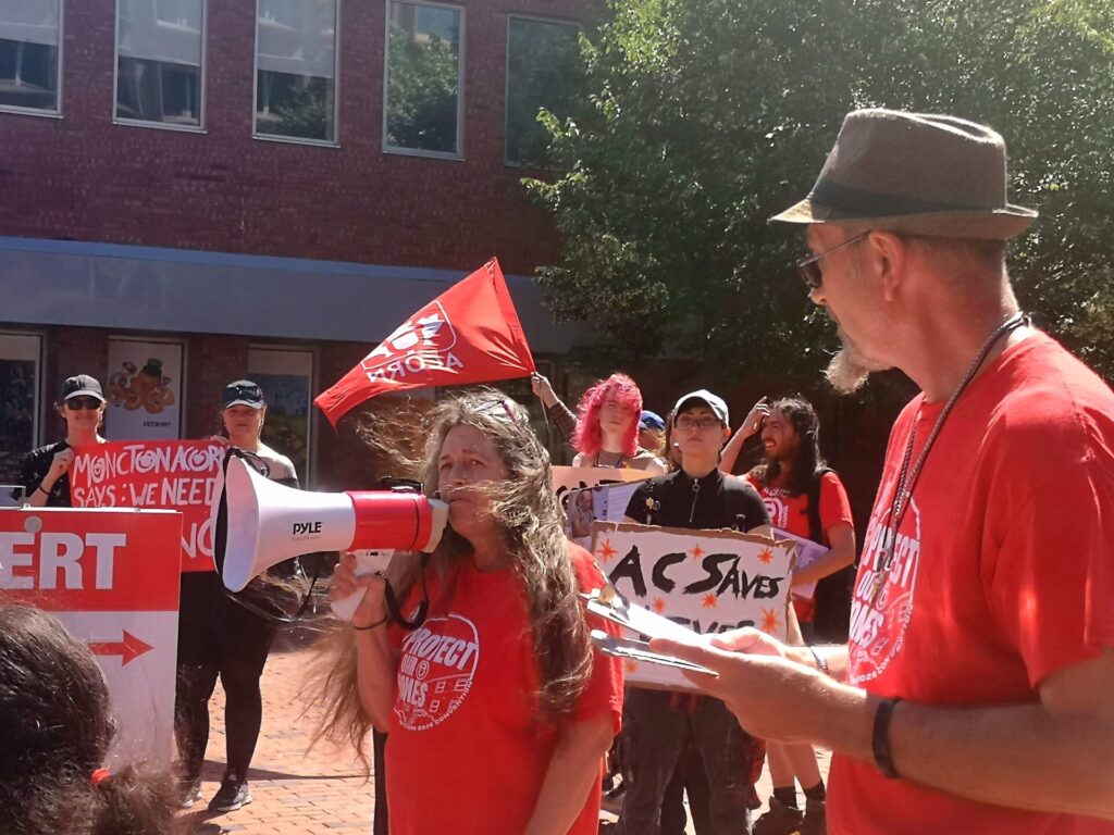 Group of protesters wearing red shirts, some holding signs and a red banner; a woman speaks into a megaphone while others hold a red flag.
