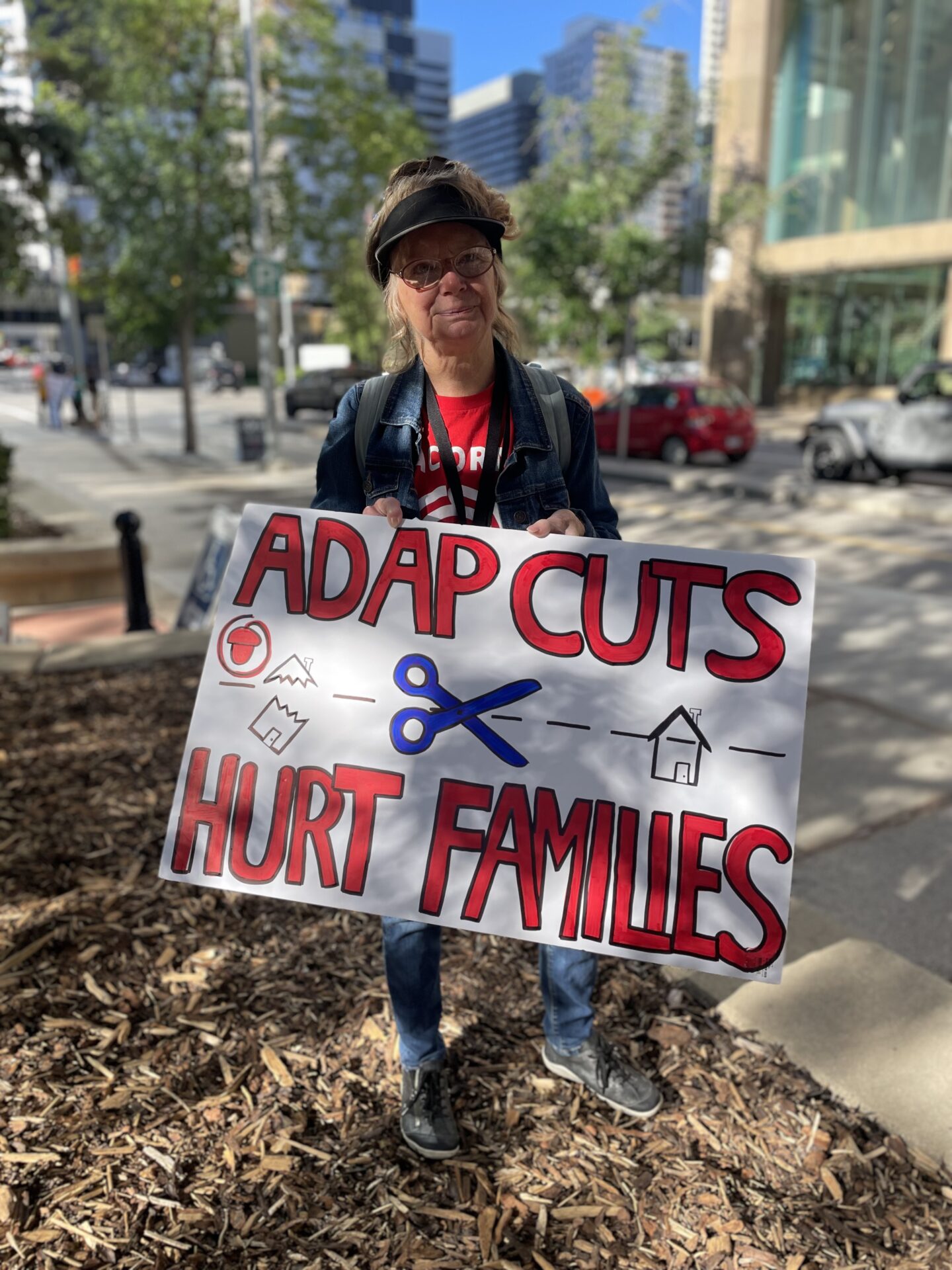 Older woman protester holding a sign that reads 'ADAPT CUTS HURT FAMILIES' on a city sidewalk, wearing a denim jacket and visor, with trees and buildings in the background.
