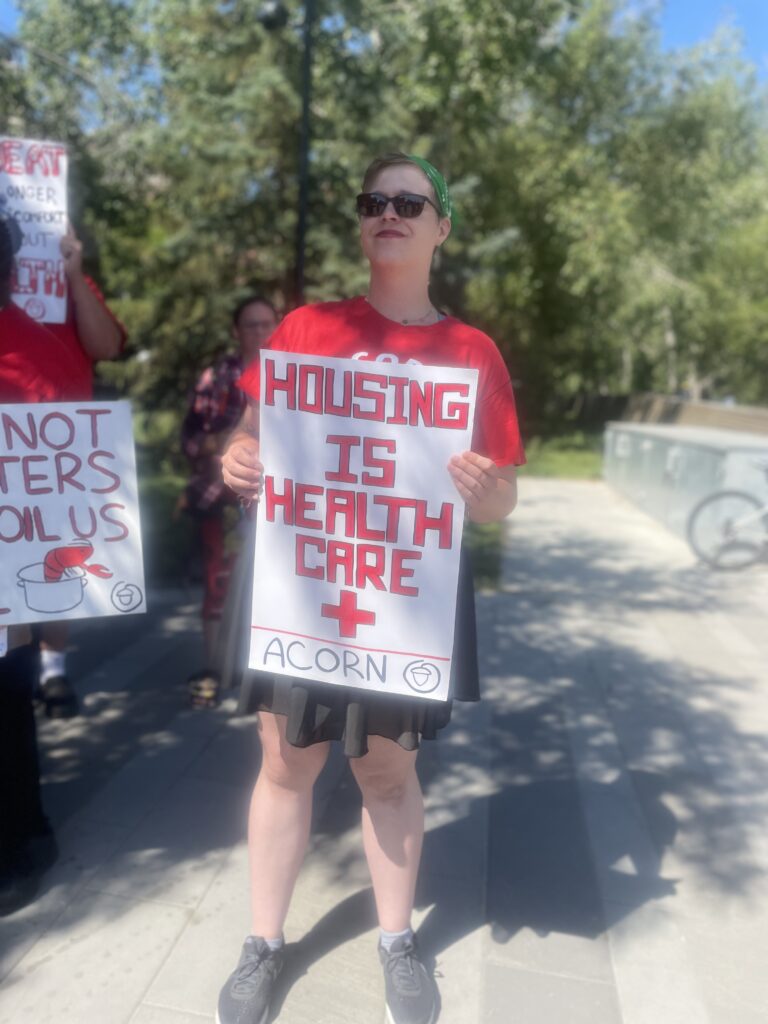 Protester in a red shirt and sunglasses holds a sign reading 'HOUSING IS HEALTH CARE' at an outdoor rally.