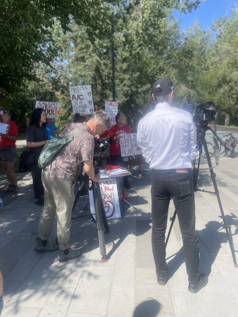A journalist in a white shirt films a protest as protesters hold handwritten signs outdoors.