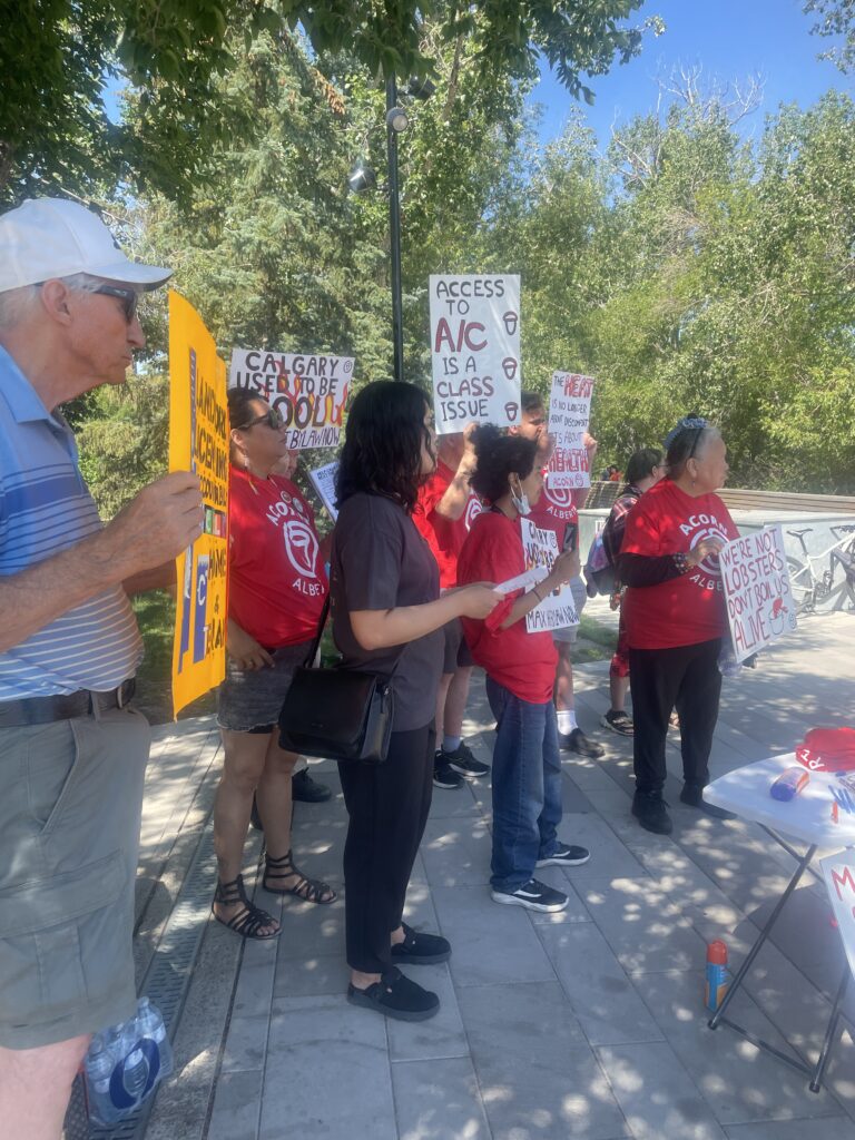 Group of protesters wearing red shirts holding signs at an outdoor demonstration under trees on a sunny day; central sign reads 'ACCESS TO A/C IS A CLASS ISSUE'.