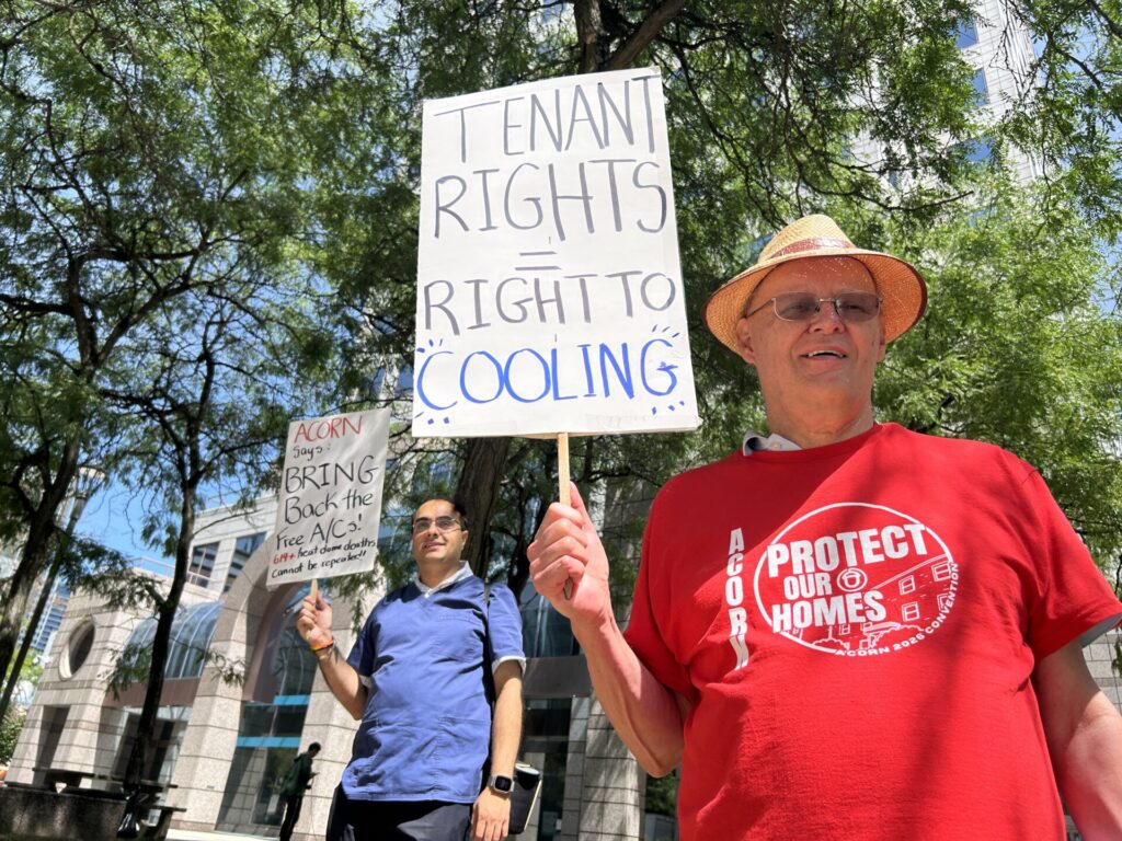 Protester in a red shirt and straw hat holds a sign that reads 'Tenant Rights = Right to Cooling' at a sunny urban protest, with another marcher and trees in the background.