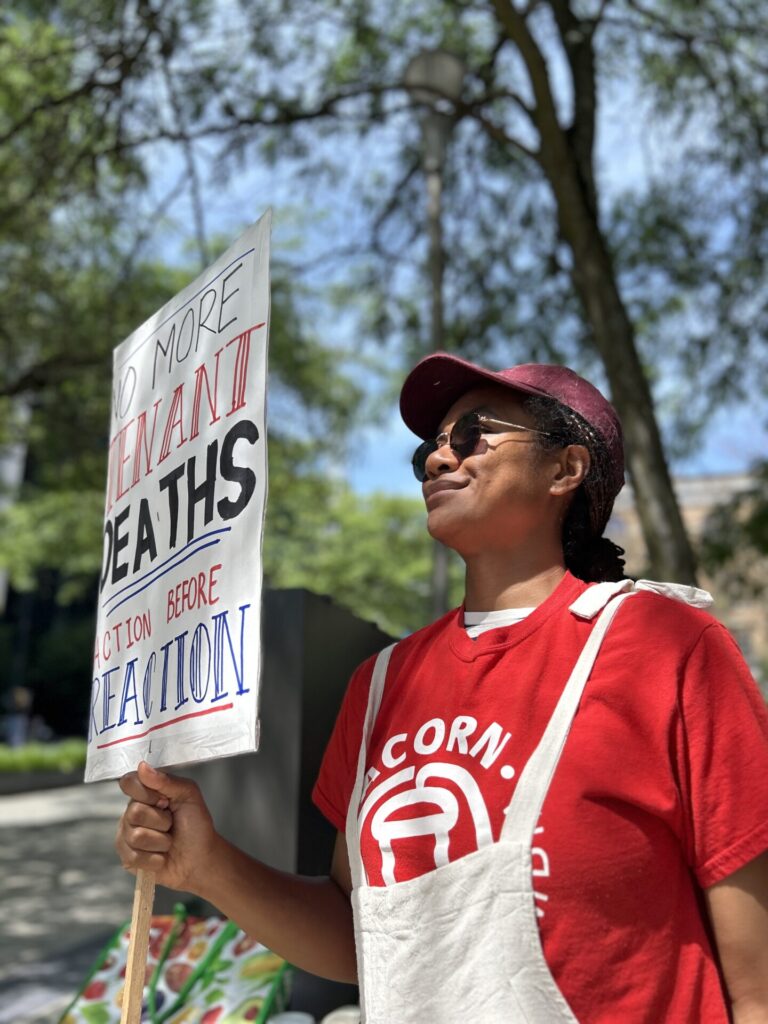 Protester in red shirt and white apron holds a sign with bold text about deaths and reactions outdoors, with trees in the background.
