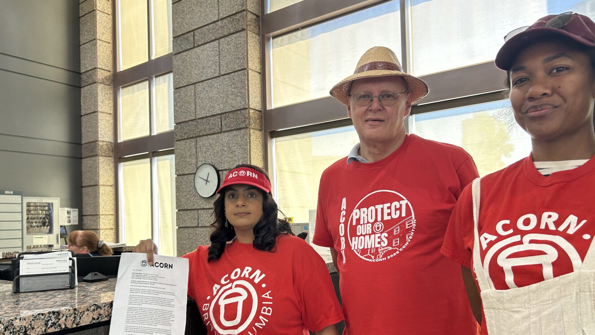 Three volunteers in red Acorn shirts at a front desk inside a building; a woman holds a clipboard and a man wears a straw hat.