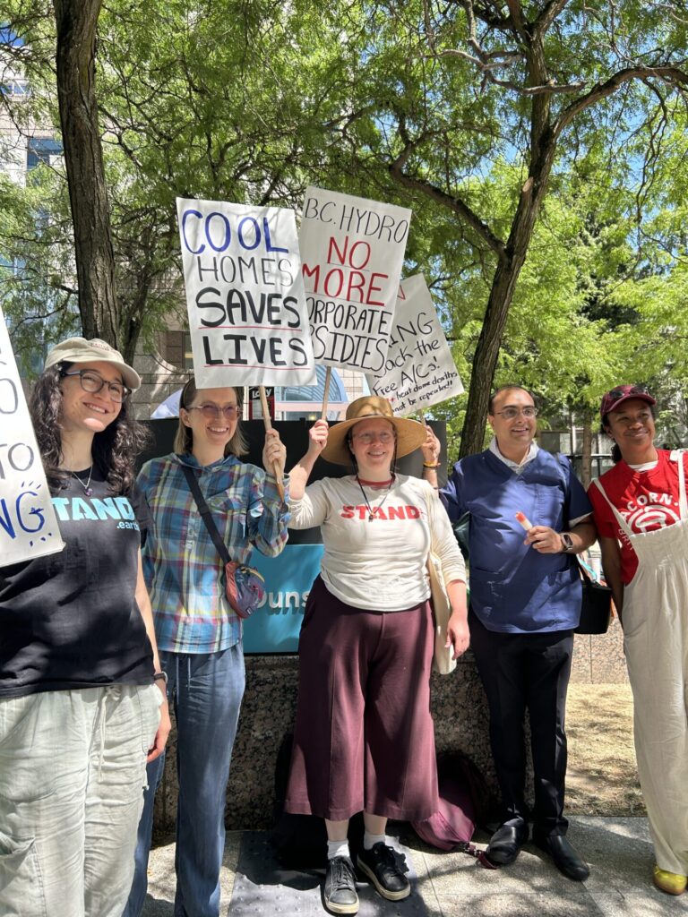 Group of protesters holding signs advocating for climate action and housing justice, outdoors under trees, with bright daylight. Sign reads 'COOL HOMES SAVES LIVES'. Other signs mention 'NO MORE ...' and 'Stand' slogans in the background.