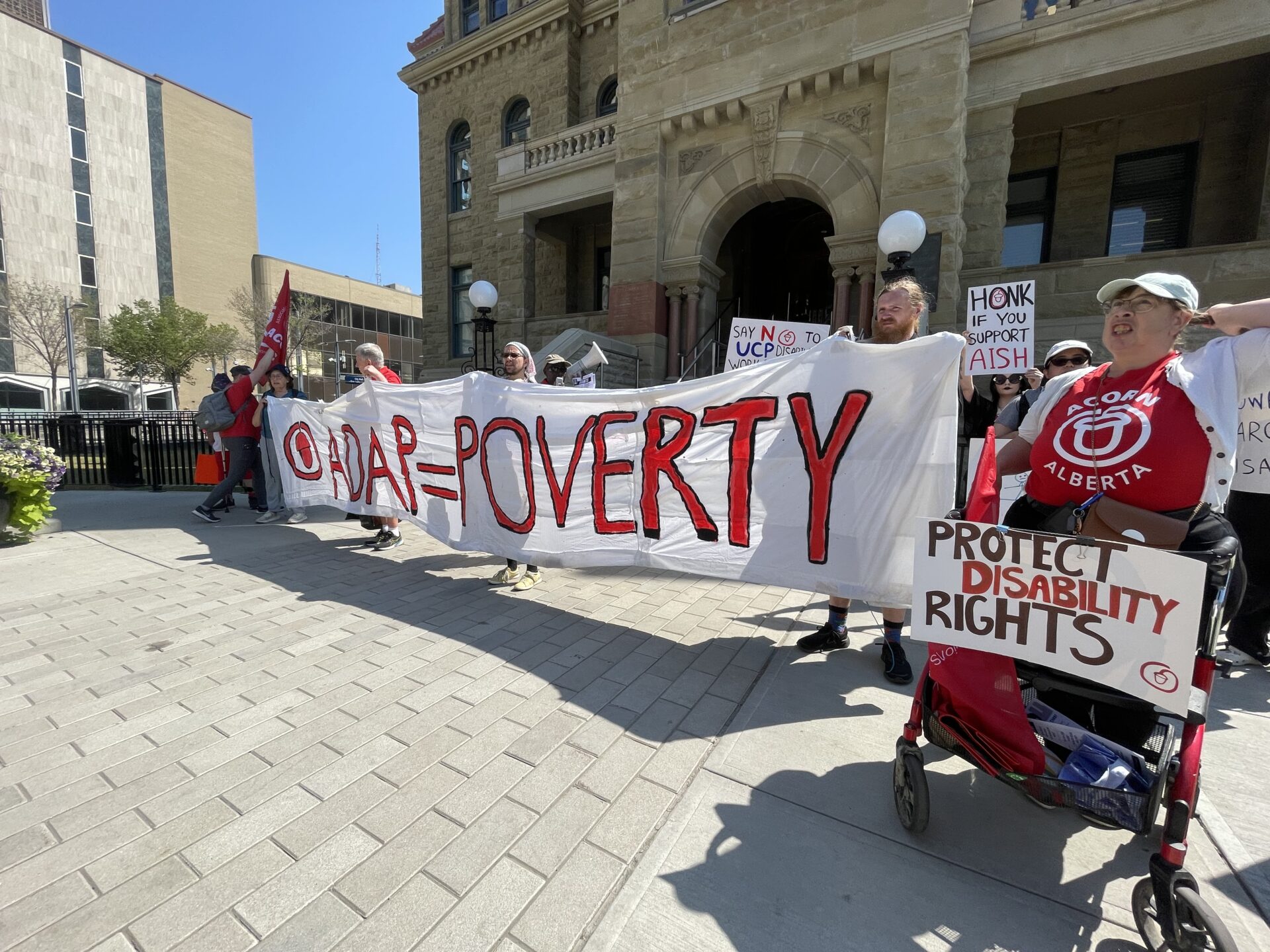 Protesters hold a large white banner with red letters reading 'POVERTY' in front of a historic stone building.