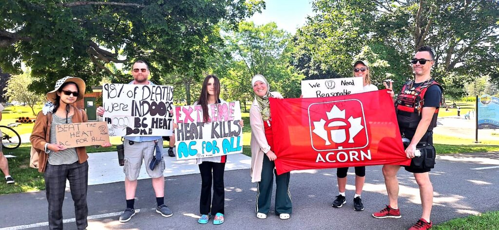 Group of six people standing on a park path, holding protest signs about heat and AC for all, with a red Acorn banner.