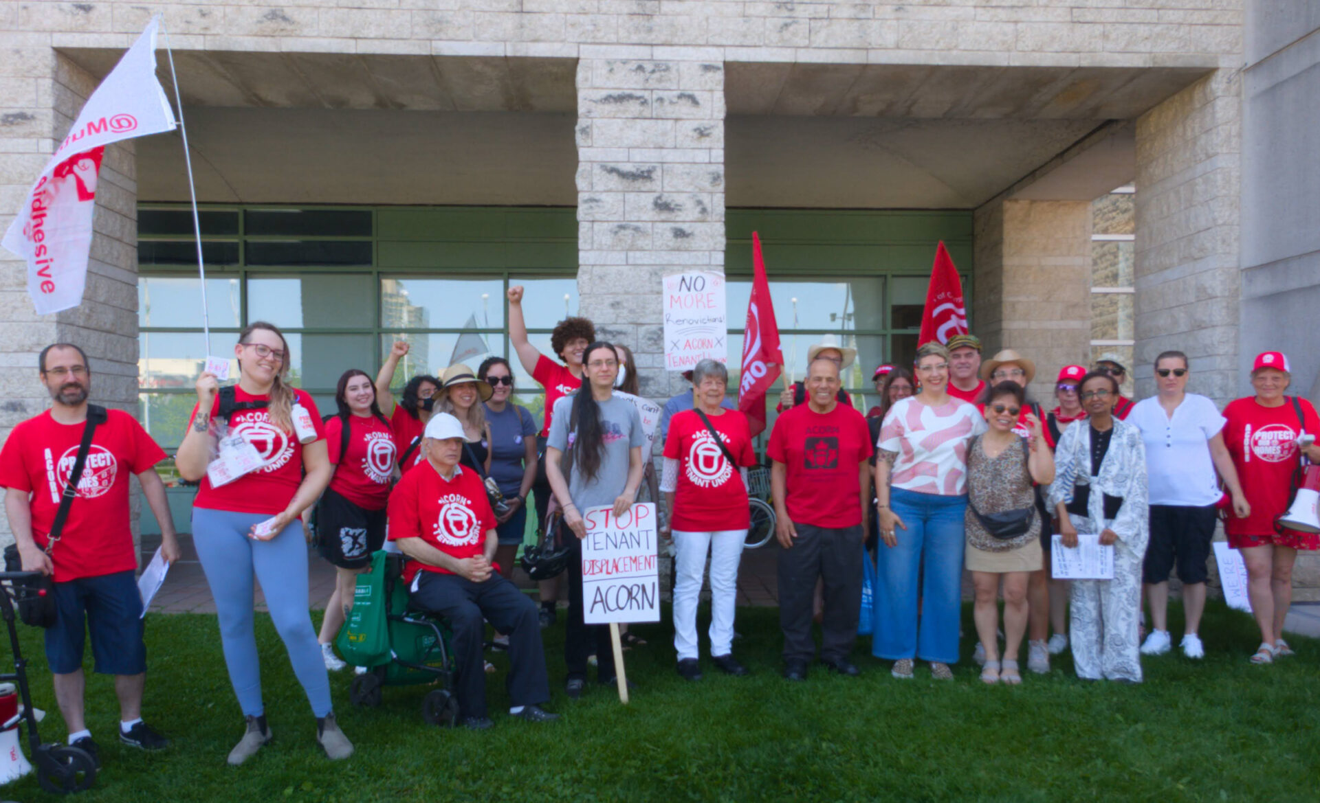 Group of diverse protesters in red shirts with signs and flags outside a stone building, raising fists in a rally feel.