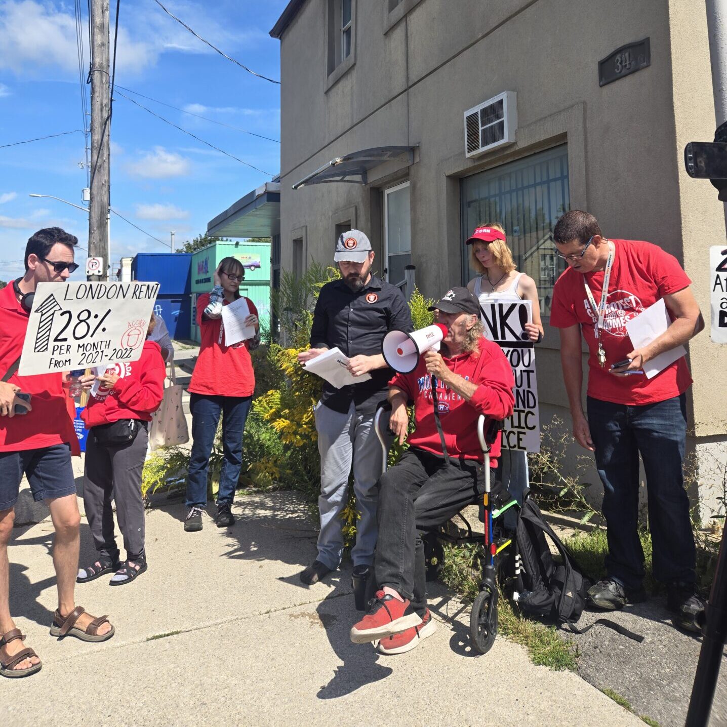 Group of protesters in red shirts on a sunny sidewalk, holding signs about London rent increases and housing justice.