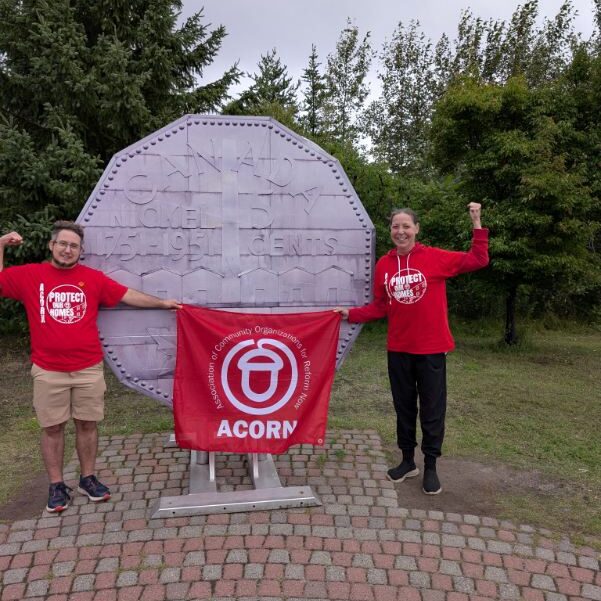 Two volunteers in red shirts pose with an ACORN banner in front of a large coin-shaped monument in a park park setting.