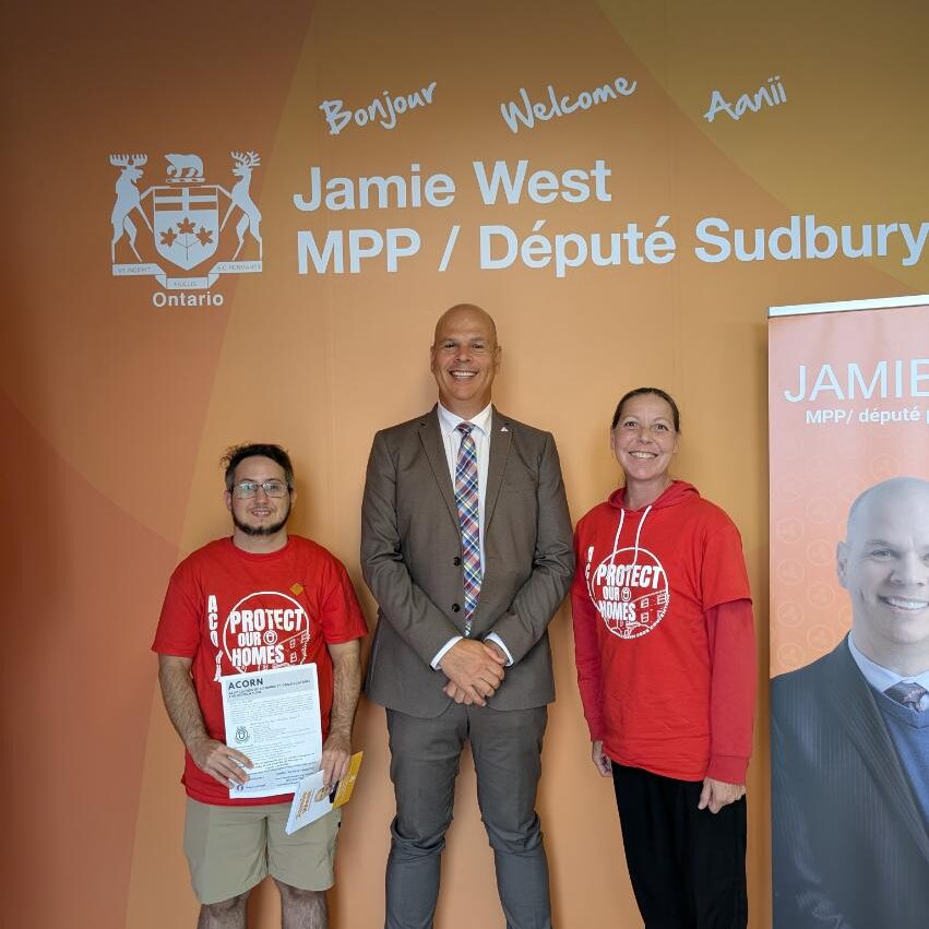 Three people pose for a photo at an Ontario campaign backdrop reading Jamie West MPP / Député Sudbury; center man in a suit, two volunteers in red shirts with a 'Protect Our Homes' logo.