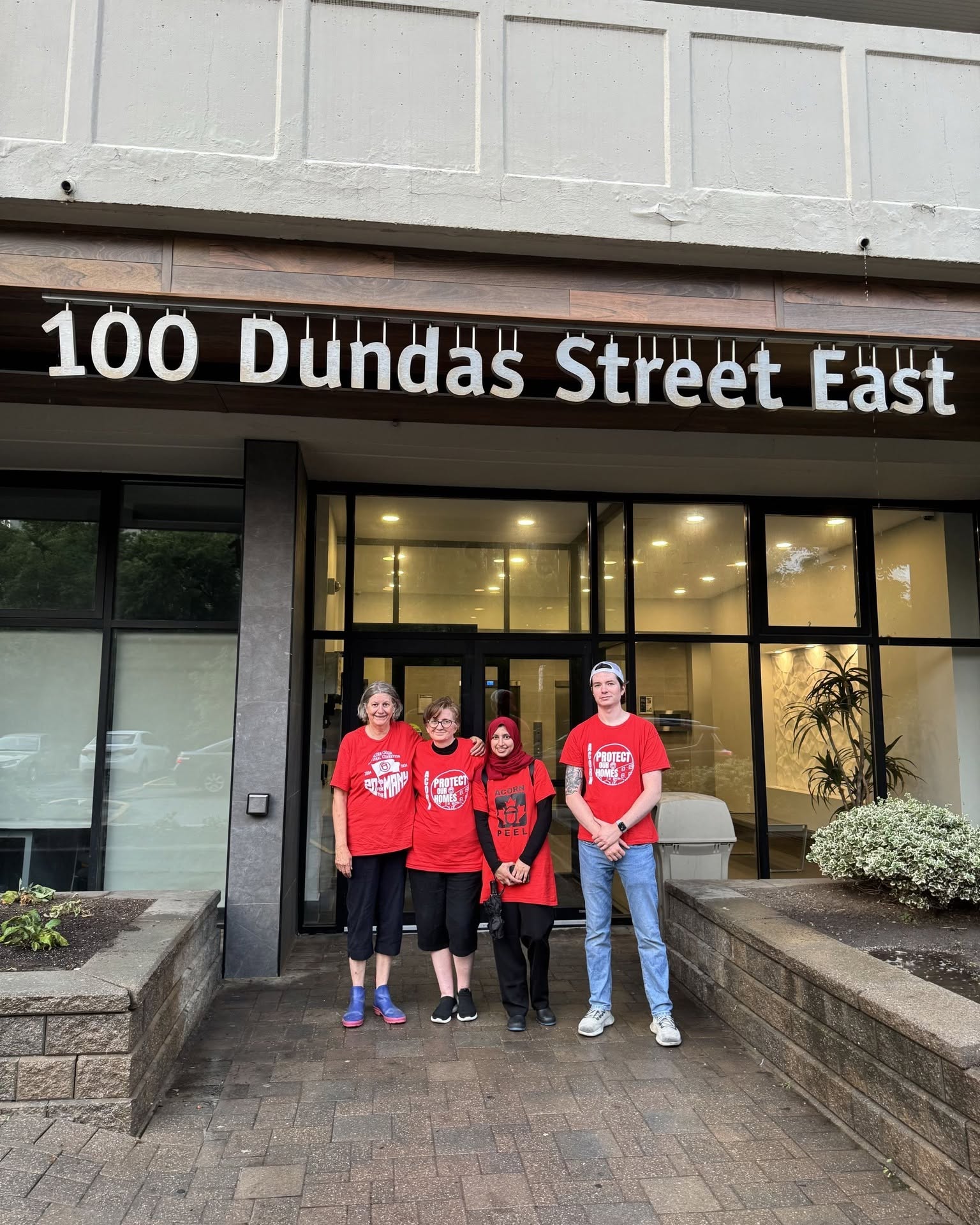 Four people in red shirts stand together outside a storefront under a sign that reads 100 Dundas Street East.