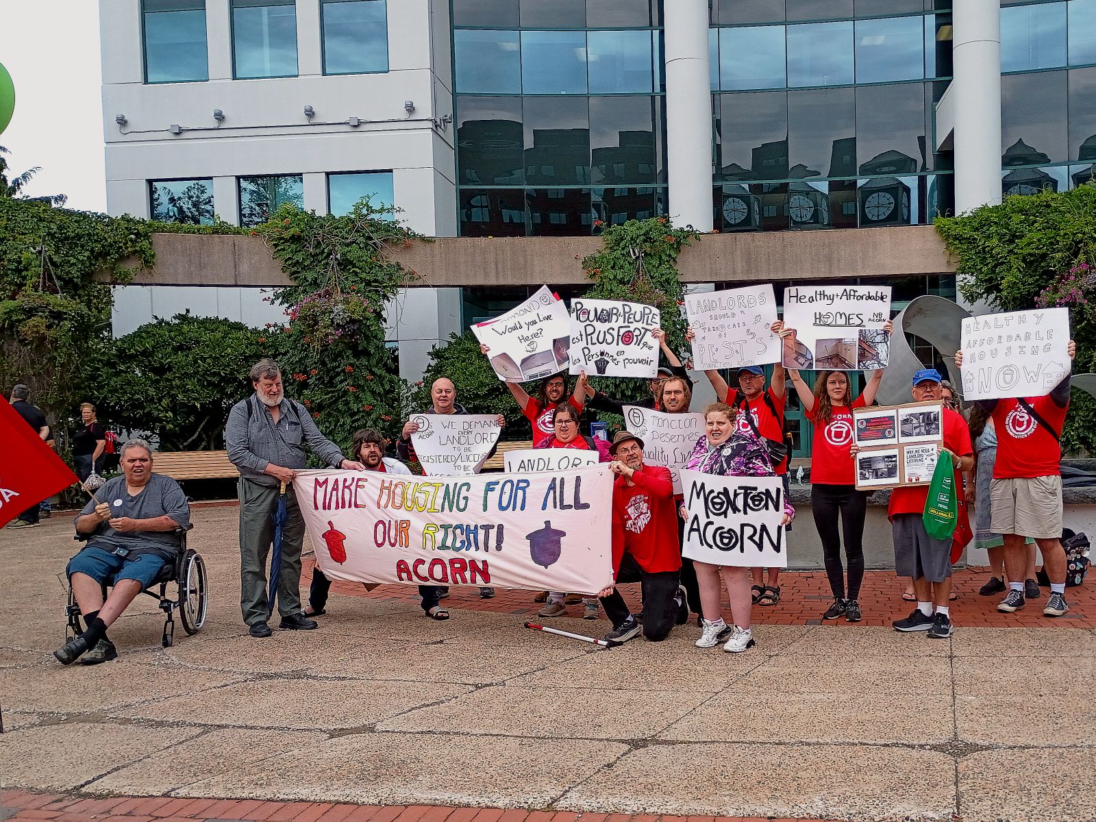 Group of protesters holding signs including 'Make Housing for All Our Right' outside a modern building; a man in a wheelchair sits on left with a red flag nearby.