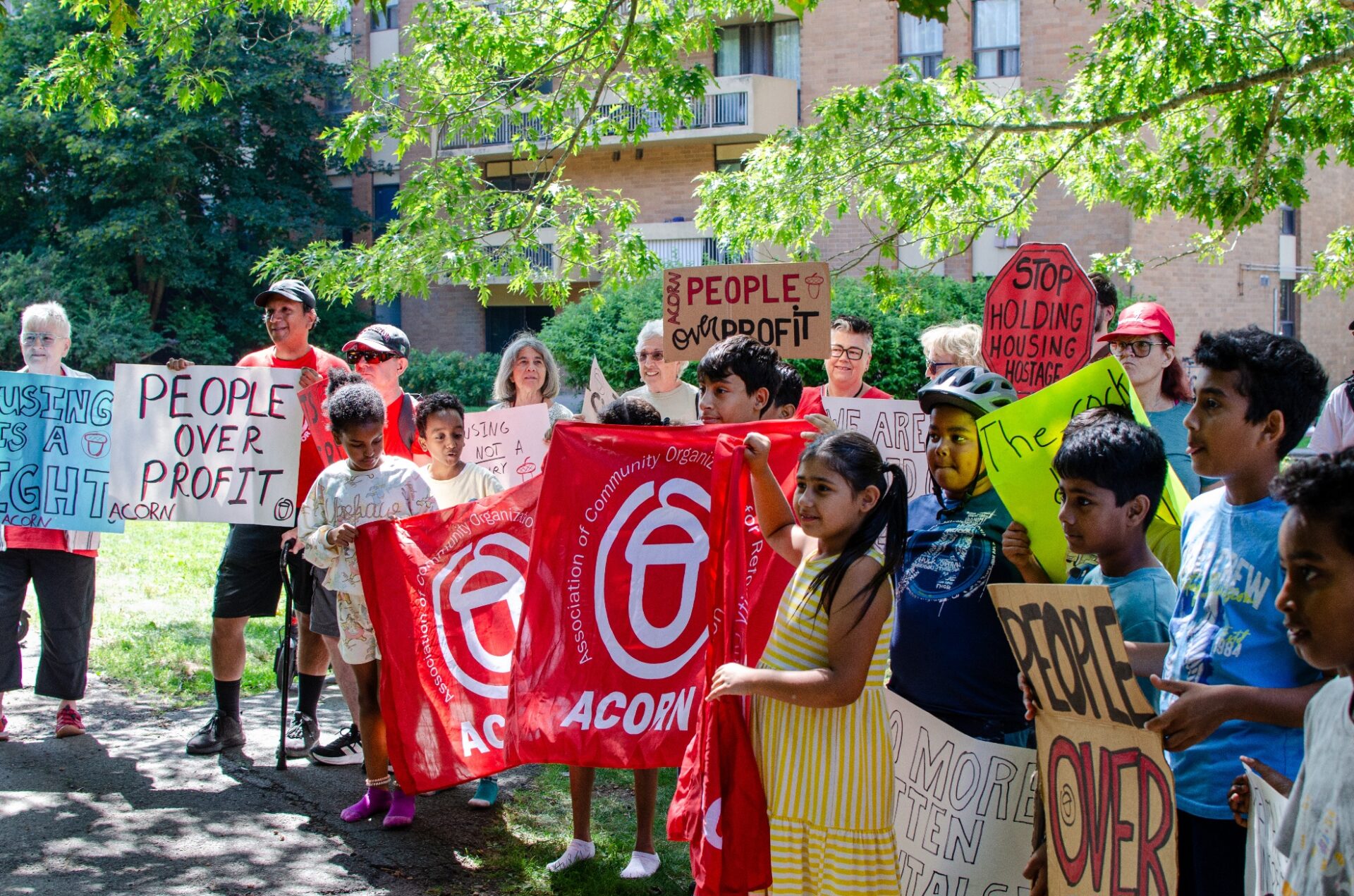 Diverse group of protesters outdoors under trees, holding handmade signs like 'People over profit' and 'Stop holding housing hostage' with red banners for ACORN.