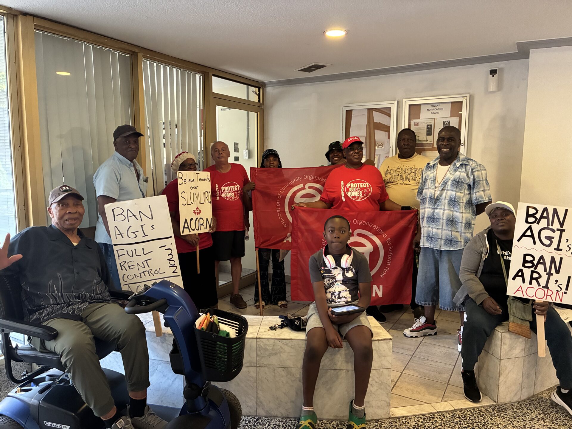 Group of tenants protesting indoors, holding signs and a red banner; several people stand together in a doorway area, including a person on a mobility scooter in the foreground. Signs partly read ‘BAN AGI’ and ‘ACORN’.