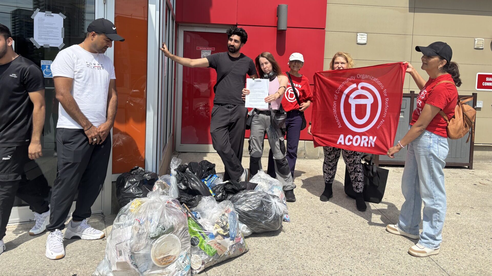 Group of volunteers posing outside a building with bags of collected trash; one person holds a red ACORN banner.