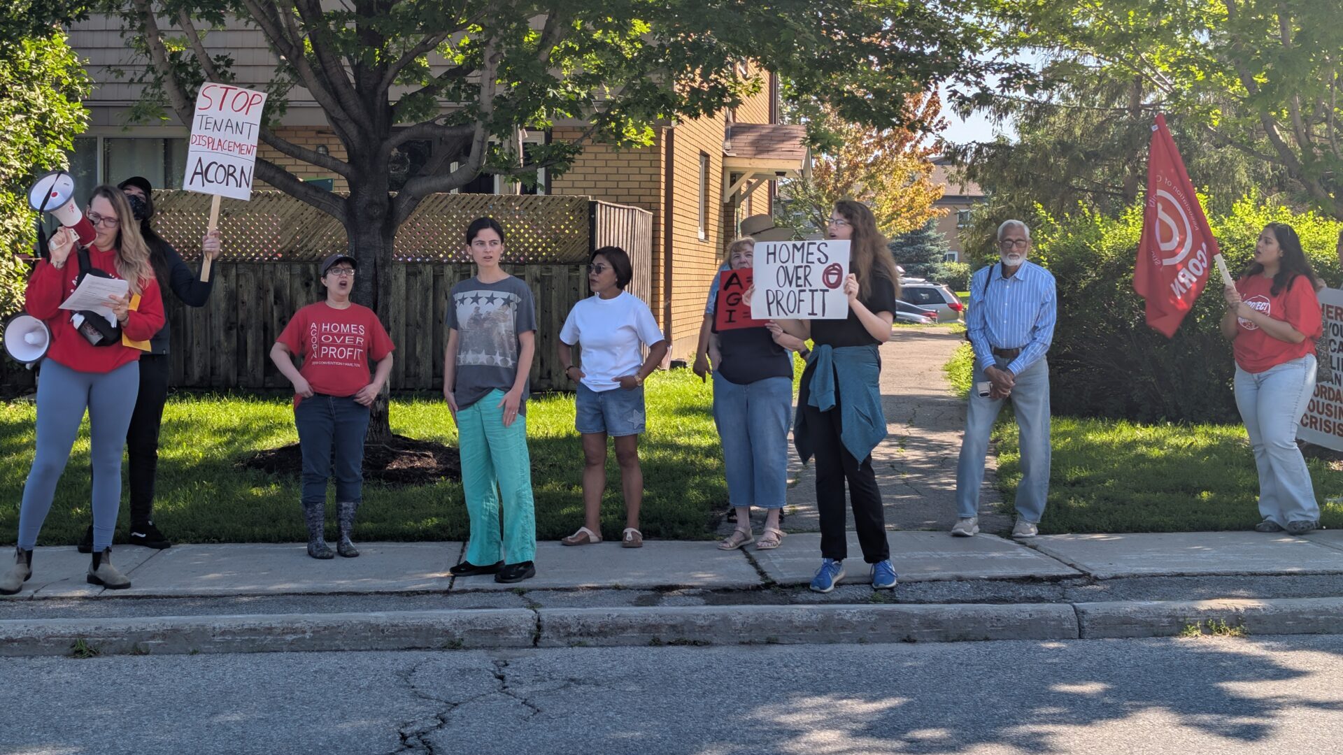 A line of protesters standing on a sidewalk holding signs that read 'STOP TENANT DISPLACEMENT' and 'HOMES OVER PROFIT' with a red flag visible on the right side. Trees and a brick house in the background.