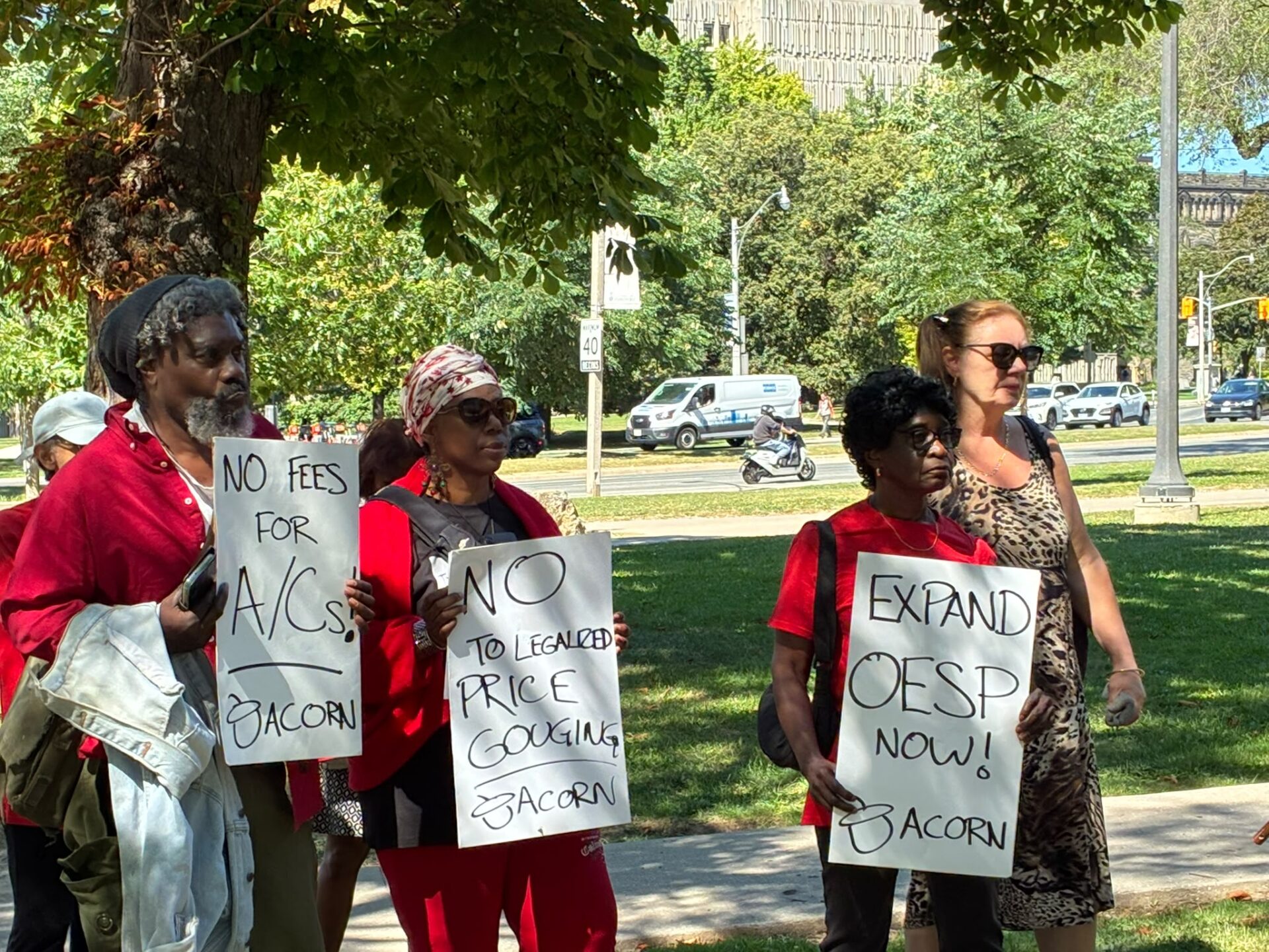 Group of diverse protesters standing in a park, each holding handwritten signs about fees and price gouging, with people wearing red clothing and sun hats.