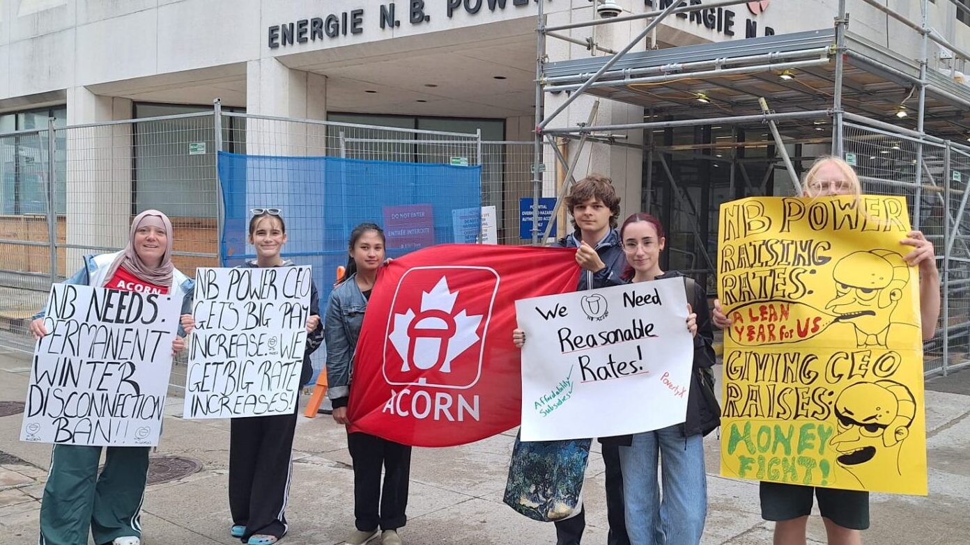 Group of young protesters outside a building with signs about NB Power, holding an ACORN banner and a yellow protest poster.
